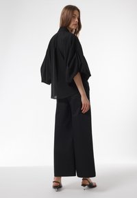 Zwarte oversized top met opgezette mouwen, gecombineerd met brede zwarte pantalons en minimalistische zwarte sandalen met puntige hielen.