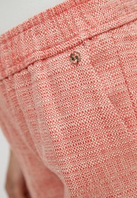 Gros plan sur un pantalon en tissu texturé rose et blanc avec une ceinture élastique et un petit ornement rond doré près de la poche.