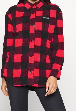 Vrouw draagt een rood-zwart geruite fleece jas met knopen en Columbia-logo op de borst, gecombineerd met zwarte legging.