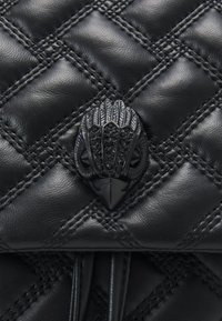 Pikowana czarna skórzana torba z błyszczącym zapięciem w kształcie ptaka, z czarnymi akcentami i szczegółowym wzorem przypominającym pióra. Teksturowana powierzchnia i eleganckie linie.