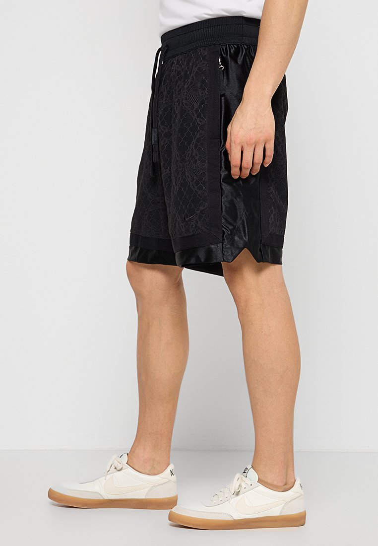 Homme portant un short de sport noir à motifs et des baskets blanches avec des semelles en gomme, se tenant devant un fond clair.