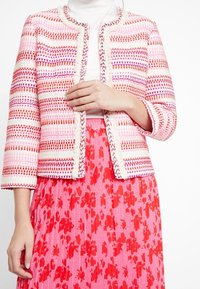 Kobieta w różowo-pomarańczowo-białej kurtce z wzorem, na białym topie i plisowanej różowej spódnicy z czerwonym kwiatowym wzorem.