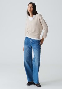 Beige gerippter Strickpullover mit weitem V-Ausschnitt, über einem weißen Hemd getragen, kombiniert mit hellblauen weit geschnittenen Jeans und schwarzen Knöchelstiefeln.