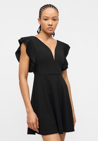 Jonge vrouw met gevlochten haar, gekleed in een zwarte jurk met geplooide korte mouwen en een diep V-hals, staand tegen een effen achtergrond.