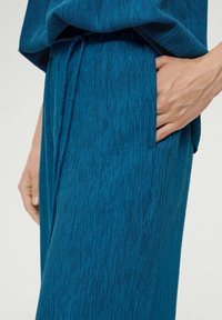 Teal textiel met een gestructureerd oppervlak en verticale groeven, met een tailleband en zijzak. Close-up van de hand die op het kledingstuk rust.