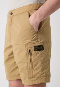 Pantaloni scurți bej dintr-un material texturat, ușor. Prezintă o buzunar frontal și un buzunar lateral cu fermoar, având un eticheta Barbour International neagră.