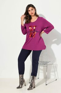 Lila Langarmshirt mit dem Aufdruck "STAY positive", kombiniert mit marineblauen Hose und metallisch silbernen Stiefeletten, vor einem neutralen Hintergrund.