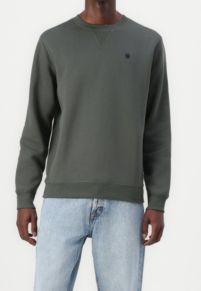 Felpa verde in cotone con scollo rotondo, polsini e orlo a coste, con un piccolo logo nero sul petto. Abbinata a jeans azzurri.