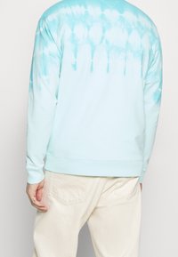 Jasnoniebieska bluza tie-dye z długimi rękawami, o miękkiej fakturze i gradientowym wzorze na plecach. Noszona z beżowymi spodniami.