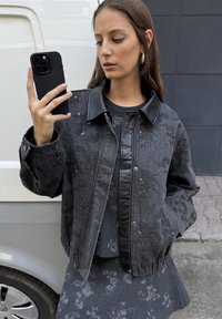 Zwarte denim bomberjack met een leren kraag, bloemenstructuur en geribde manchetten, gedragen over een bijpassende jurk met bloemenpatroon.