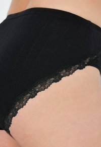 Zwarte high-waist onderbroek met verticale ribbels en een gegolfde zwarte kantafwerking langs de beenopeningen. Zachte stoftextuur.