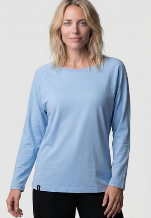 Blonde Frau, die ein hellblaues Langarmhemd und schwarze Hose trägt, vor einem einfarbigen hellen Hintergrund stehend.