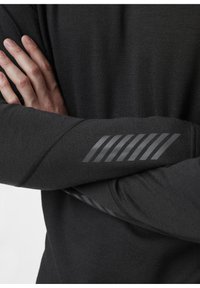 Zwart shirt met lange mouwen, gemaakt van textuurstof met een subtiele glans. De arm heeft een reflecterend gestreept patroon voor zichtbaarheid.