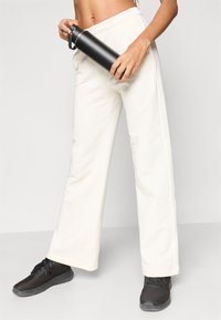 Άσπρο παντελόνι φαρδιάς γραμμής με ελαστική μέση, κρατημένο από το χέρι που κρατάει ένα μαύρο ανοξείδωτο μπουκάλι νερού, σε συνδυασμό με μαύρα αθλητικά παπούτσια.