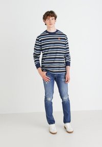 Camisa de rayas en azul marino y azul claro de manga larga con puños acanalados, combinada con unos vaqueros azul desgastados y zapatillas blancas.
