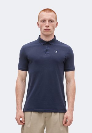 Άνδρας μοντέλο με κοντά κόκκινα μαλλιά, φορώντας μπλε ναυτικό πόλο με μικρό λευκό λογότυπο στο στήθος και μπεζ παντελόνια, στέκεται μπροστά σε απλό φόντο.