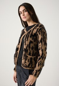 Flauschige Strickjacke in Beige mit schwarzem Tiermotiv, Knopfverschluss und Vordertaschen. Sie hat ein strukturiertes, borstiges Design.