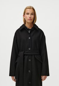 Černý vlněný kabát s předním zapínáním na knoflíky, širokým límcem a páskem v pase. Obsahuje dvě boční kapsy a elegantní, minimalistický design.