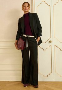 Zwarte op maat gemaakte blazer over een bordeauxrode coltrui, gecombineerd met zwarte wijde pantalon met een witte trekkoord. Houdt een bordeauxrode clutch vast.