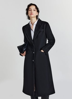 Donna che indossa un lungo cappotto nero sopra una camicia bianca, che tiene una pochette nera in una mano e con l'altra mano in una tasca.