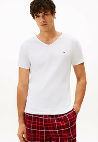 Camiseta blanca de manga corta en algodón con diseño de cuello en V. Presenta un pequeño logo en rojo y azul. Se lleva con pantalones de lounge de cuadros rojos.