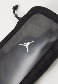 Melns aksesuāru maciņš ar caurspīdīgu priekšpusi, kurā ir balts Jumpman logo. Iekļauts melns paracorda rāvējslēdzis, kas nodrošina ērtu apstrādi.
