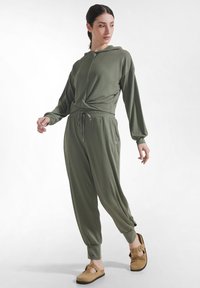 Kobieta w oliwkowozielonej, długorękawowej bluzie z kapturem i dopasowanych spodniach dresowych oraz beżowych sandałach wsuwanych, stojąca na jednolitym tle.