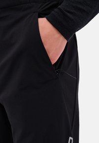 Black pants made of smooth fabric, featuring a side pocket with a drawstring, and a subtle logo detail near the hem.