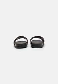 Sandales plates noires avec deux sangles croisées, une finition lisse et un design de semelle large et minimaliste. Matériau doux et flexible.