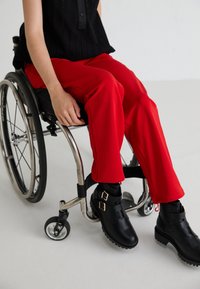 Black ankle boots with double buckles on a person in a wheelchair, wearing red tapered pants and a textured black top. Neutral background.