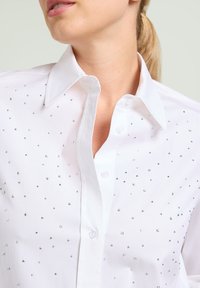 Camicia bianca con bottoni, decorata con chiodini argentati sparsi, colletto classico e una texture di tessuto liscia. I bottoni sono bianchi e semplici.