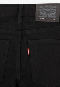 Vaqueros de mezclilla negra con bolsillos traseros con costuras curvas, una pequeña etiqueta roja y una etiqueta de marca marrón en la cintura.