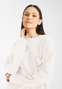 Witte gebreide trui met lichtblauwe hartpatronen, ronde hals en lange mouwen met ribboorden. Zachte textuur en een casual pasvorm.