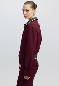 Bordeaux cropped jasje met een witte en grijze kraag, voorzien van een zijzak en elastische tailleband; gladde structuur en getailleerde pasvorm.