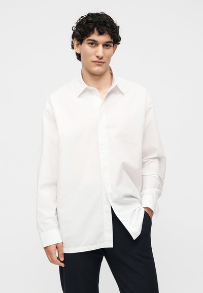 Νέος άνδρας με σγουρά σκούρα μαλλιά, φορώντας ένα φαρδύ λευκό πουκάμισο με κουμπιά και μαύρο παντελόνι σε απλό ανοιχτό φόντο.