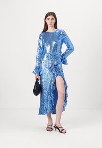 Plava haljina sa šljokicama, dugim rukavima, nabranim strukom, raširenim manžetama i bočnim prorezom. U paru s crnom torbicom i sandalama s remenčićima.