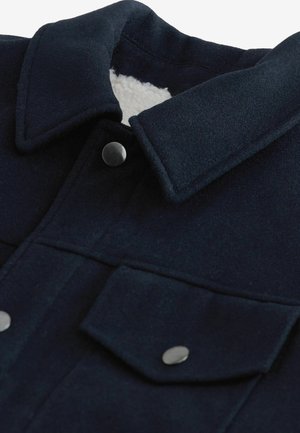 Chaqueta de lana azul marino con un interior suave y blanco, cuello puntiagudo y dos bolsillos en el pecho con botones a presión de plata.