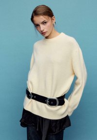 Hellcremefarbener Pullover aus weichem, strukturiertem Stoff, mit einem schwarzen Gürtel mit großer silberner Schnalle und einem geschichteten schwarzen Saum.