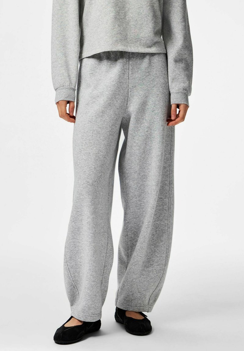 Personne portant un pantalon de survêtement gris ample, un sweat-shirt gris assorti et des chaussures noires sans lacets, debout sur un fond blanc.