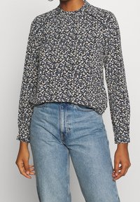 Blusa a maniche lunghe color navy con un motivo floreale in crema e giallo. Presenta un colletto alto e una vestibilità rilassata. Abbinata a jeans chiari.