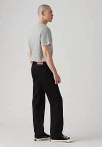 Muškarac nosi sivi majicu, crne traperice i crne patike niskog profila, stoji u neutralnoj studijskoj postavci, okrenut bočno.