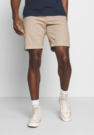 Homme portant un short beige, des baskets montantes blanches, des chaussettes blanches à la cheville et une chemise bleu marine, debout devant un fond uni.