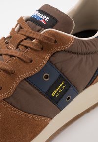 Sneaker en suède marron et nylon avec un accent bleu, une étiquette jaune « Blauer U.S.A. », des lacets texturés et une semelle en caoutchouc beige.