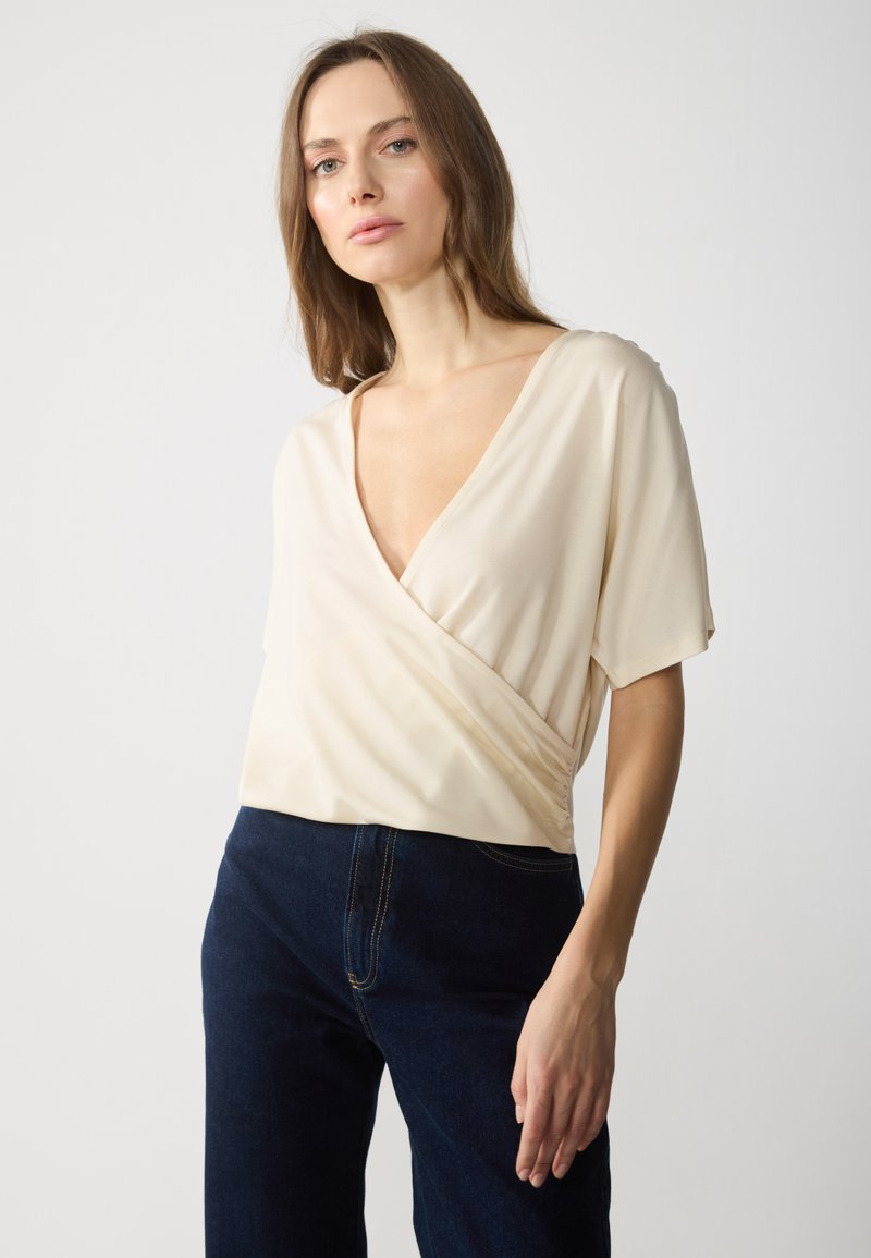 Top en beige à manches courtes, décolleté en V profond et tissu lisse. Associé à un jean bleu foncé avec des coutures contrastantes.