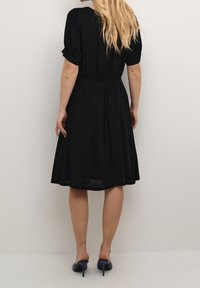 Schwarzes knielanges Kleid mit Puffärmeln, geraffter Taille und leichtem Stoff. Kombiniert mit blauen Absätzen, das Haar fällt gerade über den Rücken.