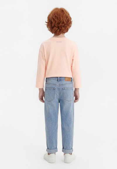 Kind mit lockigem roten Haar, das ein pfirsichfarbenes langärmliges Hemd, hellblaue hochgekrempelte Jeans und weiße Turnschuhe trägt, steht mit dem Rücken zur Kamera.