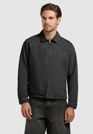 Man wearing a black zip-up jacket with collar and long sleeves, paired with dark jeans, standing against a plain light background.