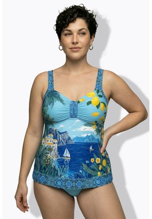 Kobieta o krótkich kręconych włosach, ubrana w niebieski tankini z żaglówkami, nadmorskimi klifami, żółtymi cytrynami i kwiatowymi wzorami, stojąca z ręką na biodrze.