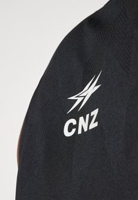 Black fabric with a textured pattern; features white "CNZ" logo and stylized graphic design. Close-up of garment material and branding detail.