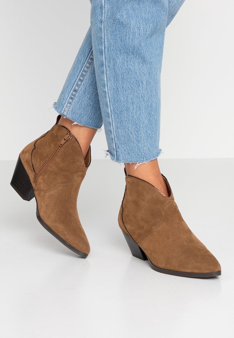 Bottes cheville en daim marron avec talons empilés portées avec un jean bleu clair à ourlet effiloché et raccourci sur un fond blanc.